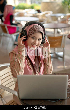Jolie jeune femme musulmane asiatique en foulard à l'écoute audio avec prise casque tout en travaillant sur laptop in cafe Banque D'Images