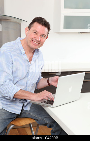 Cheerful man sitting in the kitchen en utilisant son ordinateur portable Banque D'Images