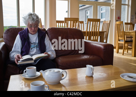 Hauts femme assise seule sur un fauteuil confortable de lire un livre Braewick café avec des tasses de thé et un verre sur une table à l'avant. UK Ecosse Shetland Banque D'Images