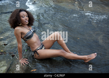 Jolie femme noire portant un maillot et assis dans un ruisseau qui coule sur la roche sombre. Banque D'Images