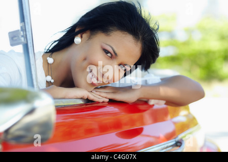 Jeune adulte brunette woman leaning on convertible voiture rouge et à la recherche à l'appareil photo. De forme horizontale, side view Banque D'Images