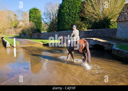 Un cheval et cavalier crossing ford dans jolie village de Cotswold Shilton, Oxfordshire, Angleterre, Royaume-Uni le soleil du printemps Banque D'Images