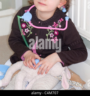 3 ans, fille, vêtu d'un cardigan brun assis se faisant passer pour un médecin avec un stéthoscope et un jouet lapin. Banque D'Images