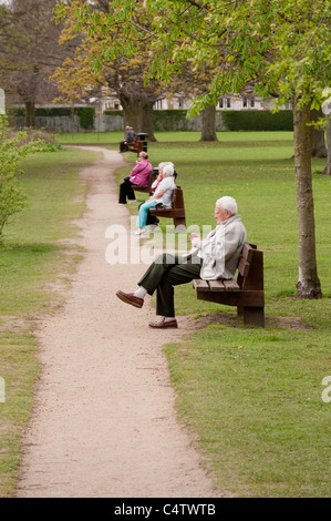 Personnes (femmes âgées et homme seul) assis se relaxant bavardant sur la ligne de bancs par chemin dans le parc pittoresque - Ilkley, West Yorkshire, Angleterre, Royaume-Uni. Banque D'Images