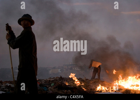 Un travailleur protège son visage de la combustion des déchets comme un autre homme est debout par à un dépotoir toxique et polluée au Cambodge. Banque D'Images