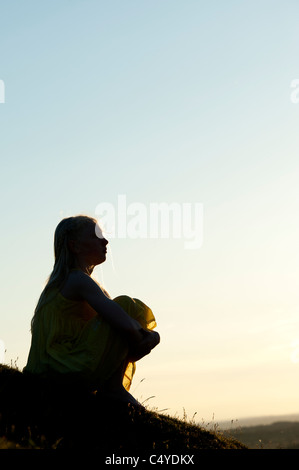 Une jeune fille assise sur une colline au coucher du soleil. Silhouette Banque D'Images