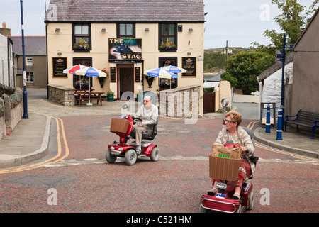 Deux personnes âgées personnes âgées équitation sur des scooters de mobilité électrique traversant une rue du village. Le Nord du Pays de Galles, Royaume-Uni, Angleterre. Banque D'Images