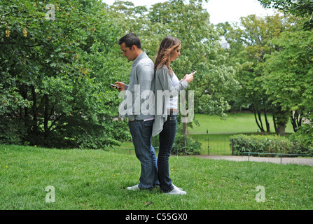 Jeune couple utilisant leur téléphone mobile dans un parc avec dos contre l'autre (posé par les modèles) Banque D'Images