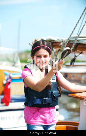 Une jeune fille portant un gilet sourit à l'appareil photo tout en maintenant des cordes sur bateau à voile sur les Norfolk Broads près de Horsey Mare Banque D'Images