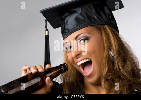 Un diplômé récent posant dans sa cap and gown holding Beer bottle isolé sur un fond argenté. Banque D'Images