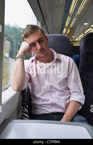 Un transport ferroviaire de voyageurs fatigués de navettage homme millénaire assis dans un siège ayant une sieste par une fenêtre voyageant sur un train quotidien England UK Banque D'Images