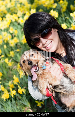 Une jeune femme tenant son chien de race mixte à l'extérieur au printemps en face d'un champ de fleurs de la jonquille. Banque D'Images