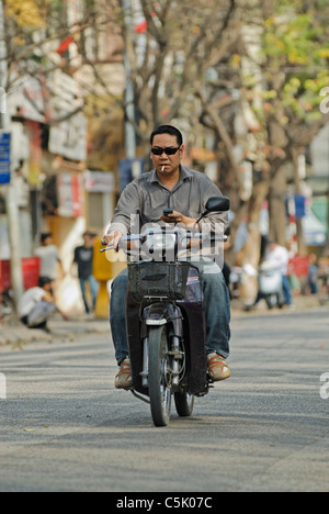 L'Asie, Vietnam, Hanoi. Vieux quartier de Hanoi. L'homme, alors que les vietnamiens cool de fumer et d'un téléphone mobile, une petite moto d'équitation Banque D'Images