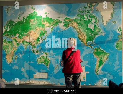 Une femme dans un manteau rouge, vue de dos, regardant une grande carte du monde sur un mur à l'aéroport d'Auckland Banque D'Images