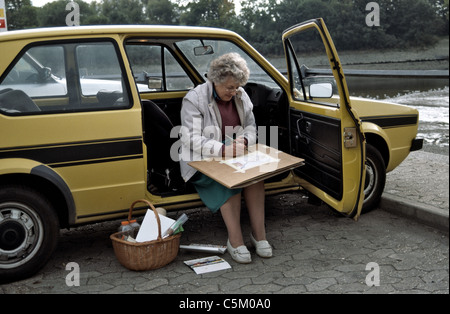 Femme âgée assise dans sa voiture avec la porte ouverte et la peinture d'une couleur de l'eau Banque D'Images