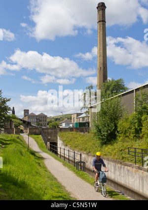 Un cycliste sur le chemin de halage du canal étroit Huddersfield avec les anciens moulins de Linthwaite, Colne Valley dans l'arrière-plan Banque D'Images