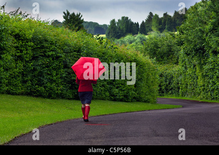 Photo d'une femme marchant sur une route tenant un parapluie rouge après une forte averse de pluie sur l'image. Banque D'Images