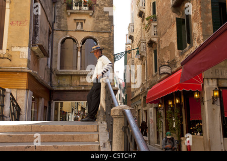 Gondolier habillé en uniforme l'attente sur un petit pont à Venise, Italie pour les clients à venir le long. Restaurants et magasins. Banque D'Images