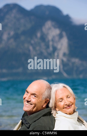 Germany, Bavaria, Senior couple relaxing on lakeshore, assis dos à dos, smiling, portrait Banque D'Images