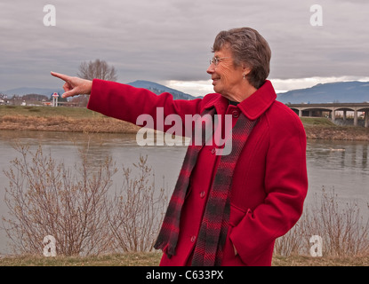 Ce portrait septuagénaire est vêtu d'un long manteau de laine rouge, à l'extérieur et en montrant quelque chose. Banque D'Images