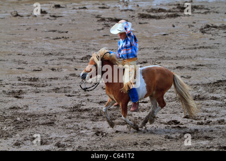 Le plus petit cowboy. Un petit garçon chevauche son poney dans l'infield boueux au Stampede de Calgary. Banque D'Images