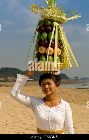 Femme balinaise en costume traditionnel marchant sur la plage portant un panier de fruits ou de canang sari offrant sur sa tête. Banque D'Images