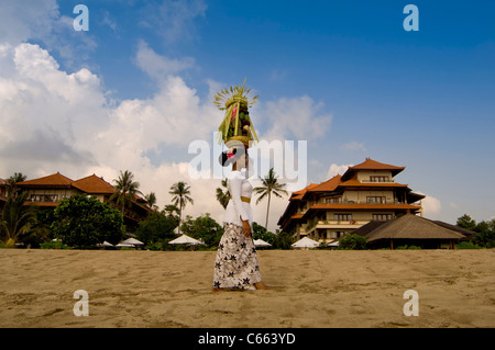 Femme balinaise en costume traditionnel marchant sur la plage portant un panier de fruits ou de canang sari offrant sur sa tête. Banque D'Images