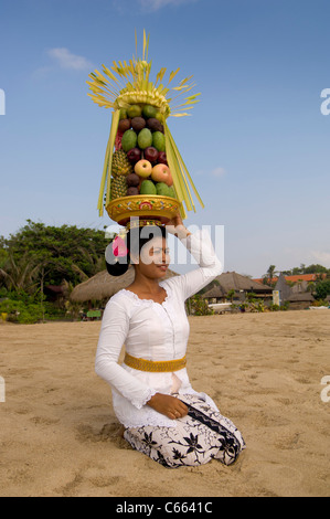 Femme balinaise en costume traditionnel s'agenouillant sur la plage portant un panier de fruits, également connu comme canang sari sur sa tête. Banque D'Images