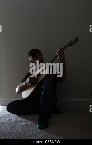 Jeune homme assis seul jouant une guitare acoustique dans une pièce sombre avec une porte ouverte. Banque D'Images