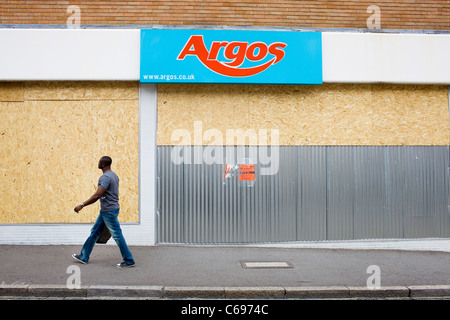 Un homme noir passe devant un magasin Argos barricadèrent endommagées dans les émeutes de Londres. Banque D'Images