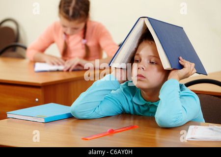 Cute schoolboy garder ouverte livre sur tête en classe Banque D'Images