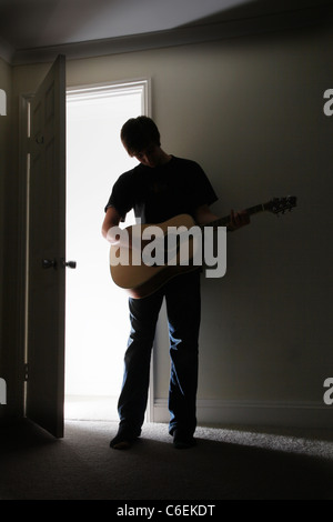 Jeune homme debout dans une pièce sombre jouant une guitare acoustique. Banque D'Images