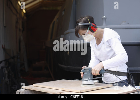 Le ponçage d'une femme une porte en bois à chevalets, porter des protège-oreilles, des lunettes, des gants en latex et un masque de protection. Banque D'Images