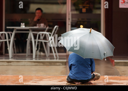 Sans-abri assis sur trottoir avec parapluie sous forte pluie en face de café bar pour demander la charité, Vientiane, Laos Banque D'Images