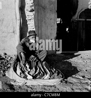 Années 1950. Photo historique montrant un homme barbu avec chapeau assis à côté de l'ancien bâtiment, la vente des couvertures, Isfahan, Iran. Banque D'Images