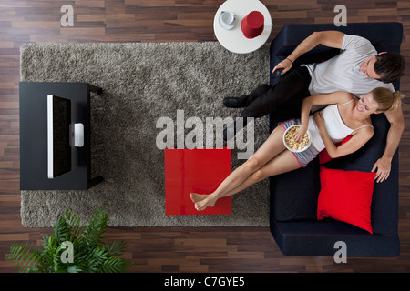 A cheerful couple watching TV and eating popcorn dans leur salon, overhead view Banque D'Images