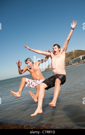 Deux jeunes hommes 2 sauts dans la mer par une chaude journée de fin septembre quand une vague frappe le Royaume-Uni Banque D'Images