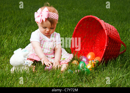 Bébé jouant avec des oeufs de Pâques tout en étant assis à côté d'un panier rouge renversé Banque D'Images