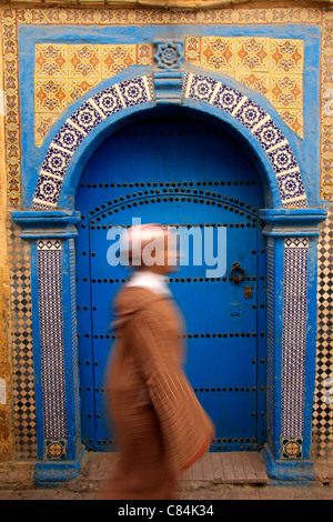 Jeune femme portant une jellabah passe devant une porte en bois et sol carrelé décoratif dans la médina/vieille ville fortifiée, Essaouira, Maroc Banque D'Images