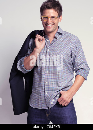 Sourire à la mode homme dans la trentaine portait un jean et une chemise avec une veste par-dessus son épaule Banque D'Images