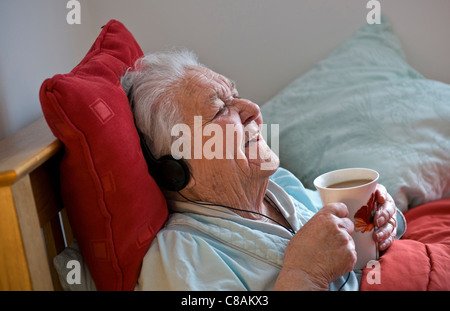 Heureux satisfait dame âgée resting in bed wearing headphones bénéficie d'une tasse de thé dans sa chambre confortable ensoleillée Banque D'Images