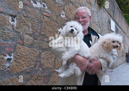 Un homme d'âge moyen détient deux petits chiens blancs dans ses bras tout en marchant dans une rue de Chianciano Terme, Ombrie, Italie, Europe. Banque D'Images