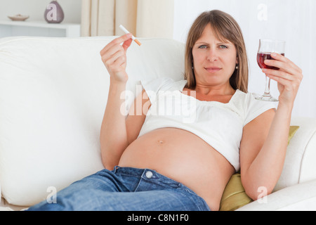 Charmante femme enceinte tenant une cigarette et un verre de vin rouge en position allongée sur un canapé Banque D'Images