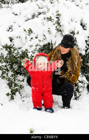 Maman joue avec 1 ans Bébé garçon dans la neige en face de bush alors qu'il neige dans la région de Hyde Park à Londres. Banque D'Images