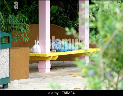 Un homme dort sur un banc dans la chaleur de la journée. Ses effets personnels en lieu sûr dans un sac en plastique, ses chaussures à ses côtés. Banque D'Images