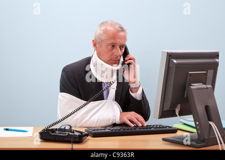 Photo d'un homme d'âge mûr avec des blessures à parler au téléphone tout en essayant de travailler sur son ordinateur. Banque D'Images