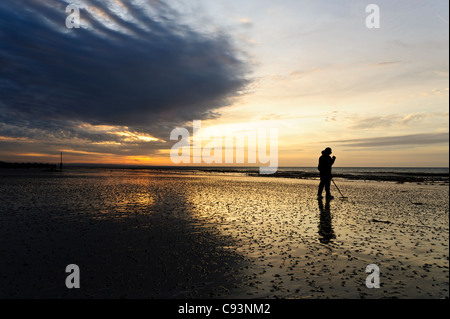 Un homme en peignant sur la plage de Worthing Beach en utilisant un détecteur de métal au lever du soleil. Banque D'Images