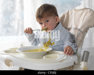 Un an et demi ans garçon assis sur une chaise haute et de manger la soupe avec une cuillère, déversant sur son T-shirt Banque D'Images