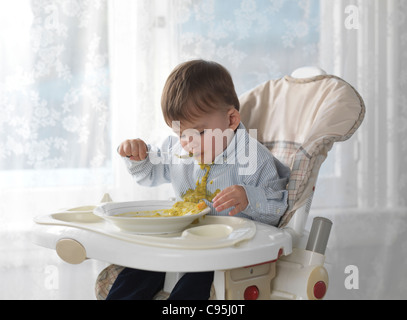 Un an et demi ans garçon assis sur une chaise haute et de manger la soupe avec une cuillère, déversant sur son T-shirt Banque D'Images