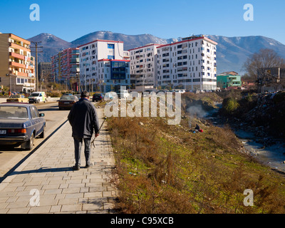 Un homme marche le long Bulevardi Zhan D'Arche, Tirana, Albanie. Au-delà, un homme fait fondre des métaux au-dessus d'un feu sur les bords de l'allée River. Banque D'Images